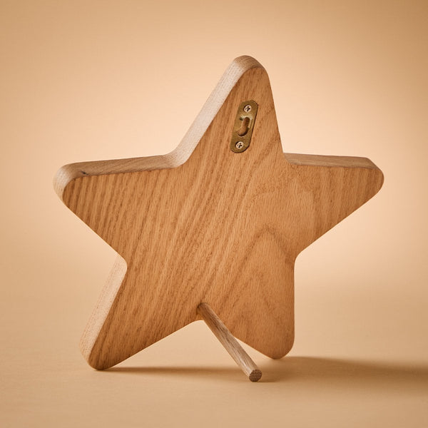 A wooden star-shaped tabletop stand with a natural oak finish and a small brass hook on the back, shown on a warm beige background.