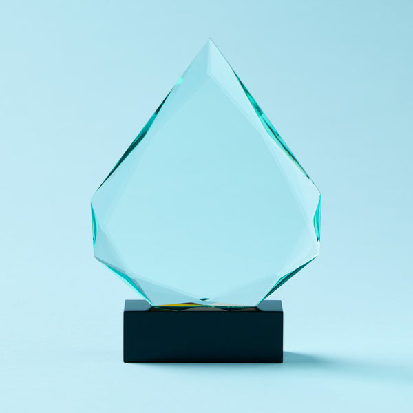 A faceted clear glass award trophy with a geometric diamond-like shape, displayed on a black rectangular base against a light blue background.