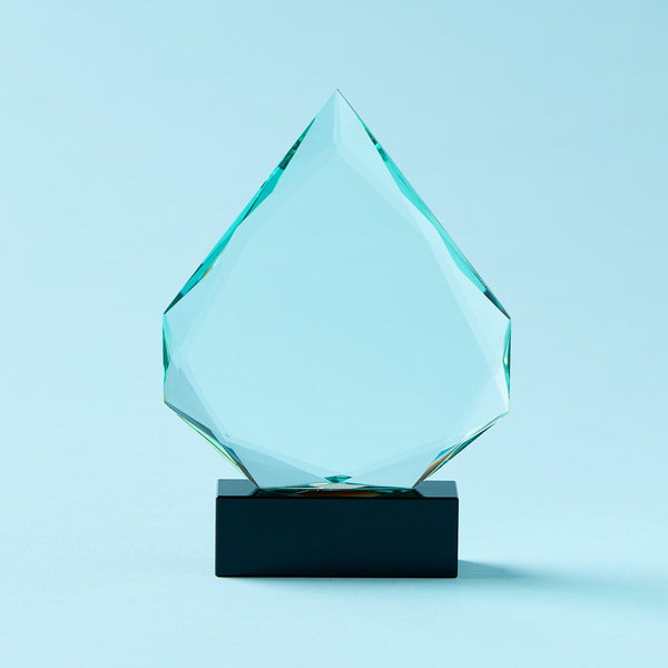 A faceted clear glass award plaque shaped like a diamond, mounted on a rectangular black base, set against a solid light blue background.