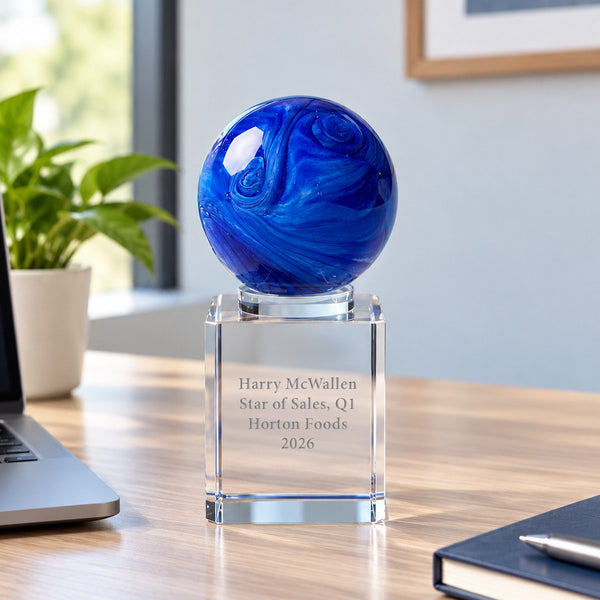 A personalized blue glass sphere award on a clear engraved crystal base, displayed on a wooden desk in an office setting.