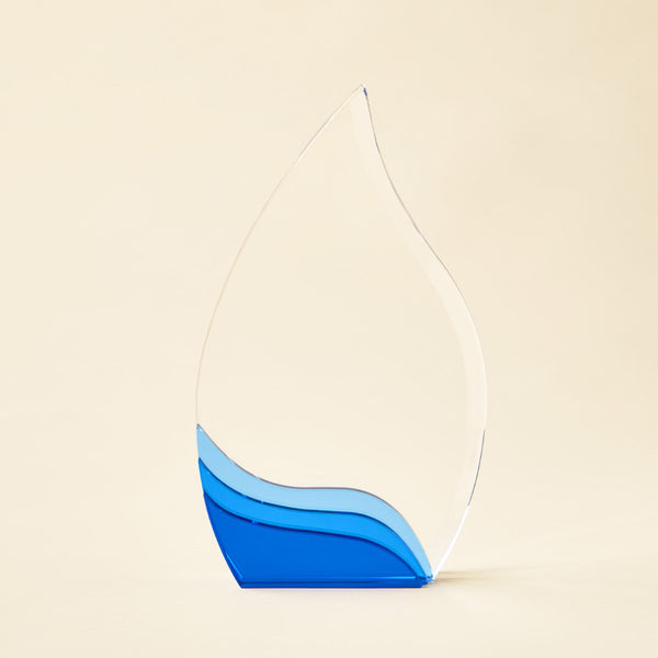 A clear glass award trophy shaped like a teardrop, featuring a layered blue wave design at the base, set against a solid light beige background.