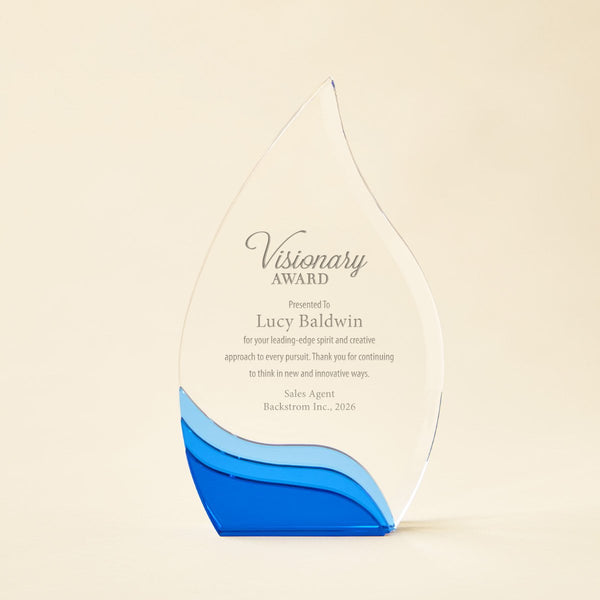 A clear glass award with a teardrop shape, featuring a blue wave design at the base and printed text reading 'Visionary Award' and 'Presented to Lucy Baldwin.' The trophy is photographed on a plain warm beige background.