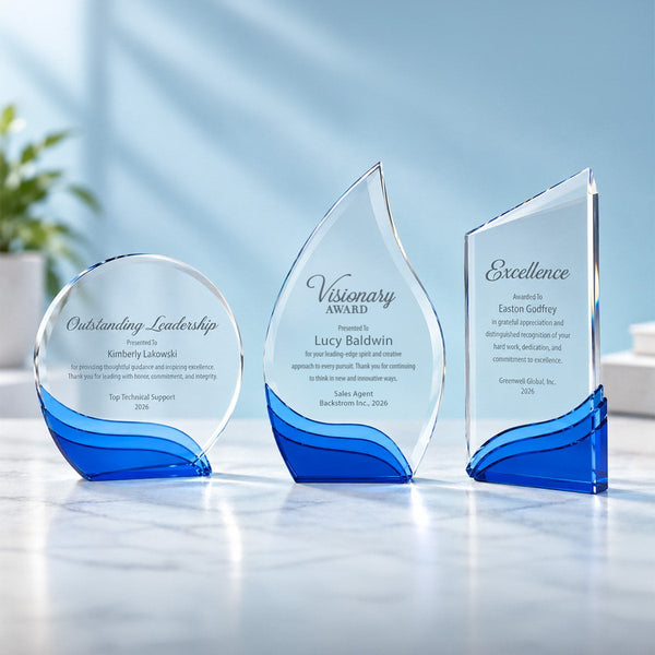 Three clear glass custom awards with vibrant blue wave-like bases, displayed on a light marble surface against a soft blue background.