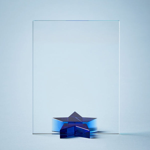 A clear crystal or glass award plaque with a small blue star-shaped pedestal at the base, photographed against a soft light blue studio background.