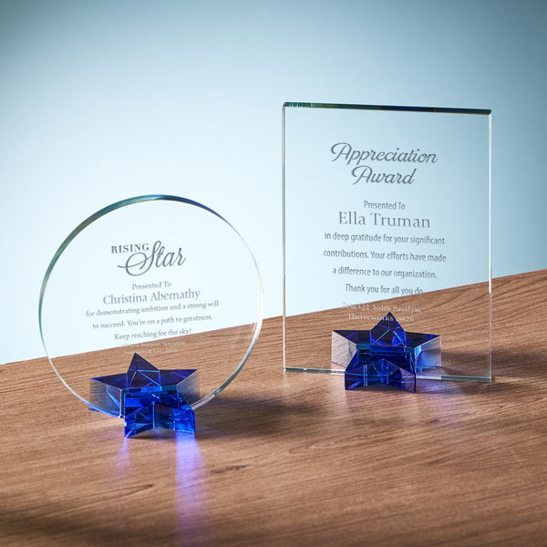 Two clear crystal award plaques on a wooden surface against a soft blue gradient background. One round plaque sits on a blue star-shaped base, and one tall rectangular plaque stands on a matching blue star base.